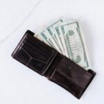 About From above of black leather wallet with pockets filled with heap of dollar banknotes on white table